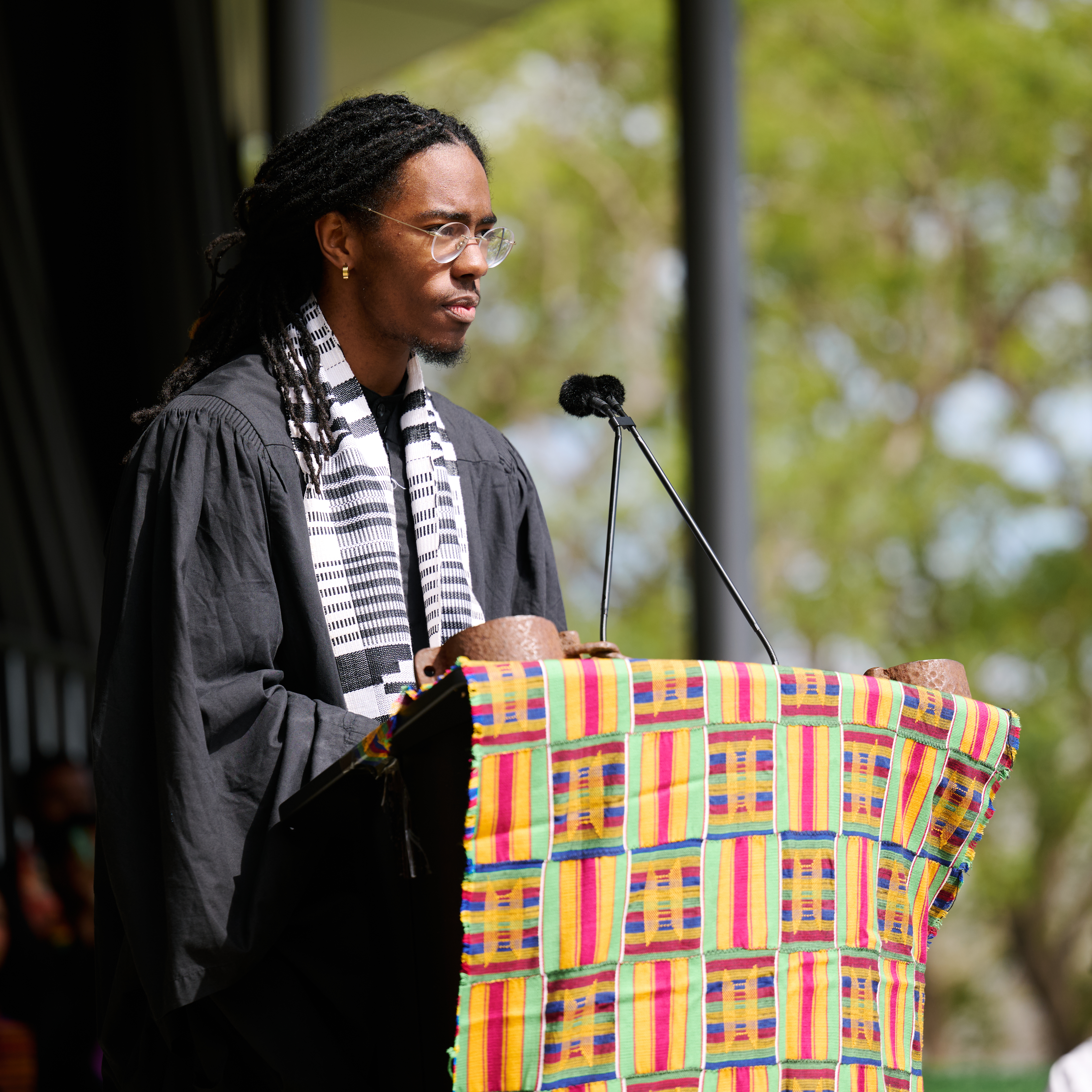 A student stands at the podium outside on Drollinger Family Stage speaking at Kente wearing a black robe.
